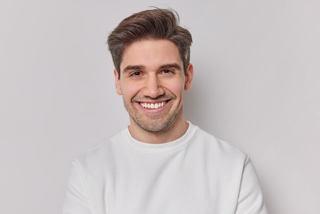 Portrait handsome man with dark hairstyle bristle and toothy smile dressed in white sweatshirt feels very glad poses indoor Pleased European guy being in good mood smiles positively Emotions concept