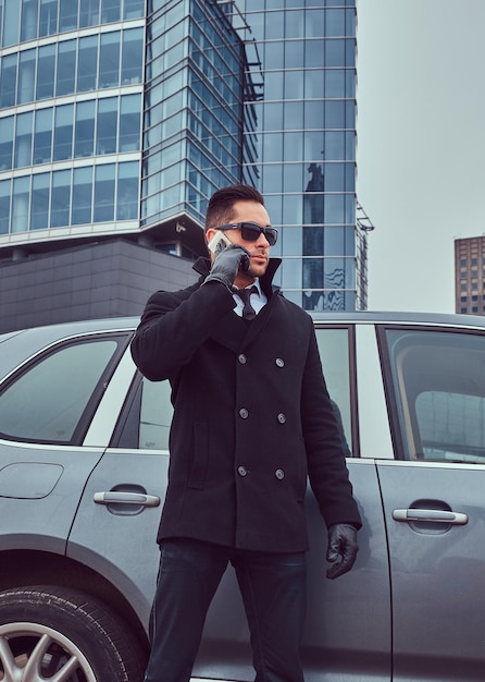 Free photo portrait of a handsome serious male bodyguard in sunglasses talking on the phone near car outdoors.