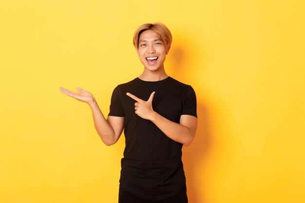 Portrait of handsome smiling asian guy holding something on hand and pointing finger at it, standing yellow wall pleased.