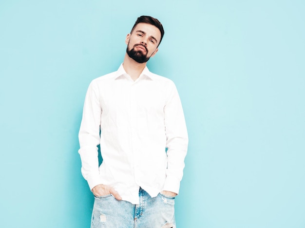 Portrait of handsome smiling model Sexy stylish man dressed in white shirt and jeans Fashion hipster male posing near blue wall in studio Isolated