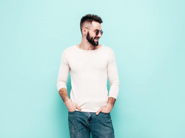 Portrait of handsome smiling stylish hipster lambersexual modelMan dressed in white sweater and jeans Fashion male posing near blue wall in studio in sunglasses Isolated