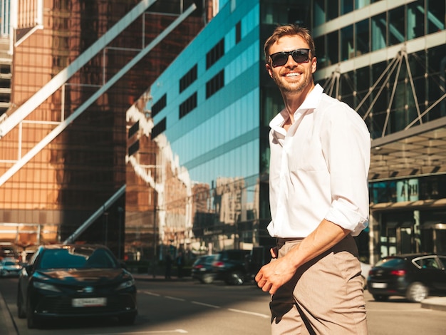 Portrait of handsome smiling stylish hipster lambersexual modelModern man dressed in white shirt Fashion male posing in the street background near skyscrapers in sunglasses