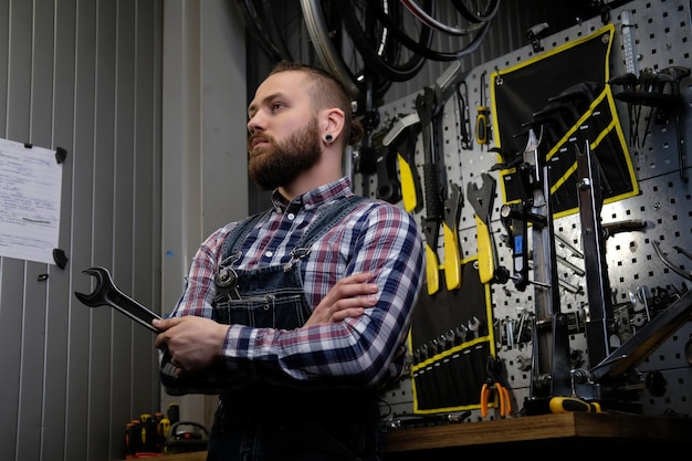 Free Photo portrait of a handsome stylish male with beard and haircut wearing a flannel shirt and jeans coverall, holds steel wrench, standing in a workshop against wall tools.