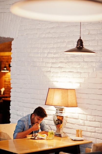 Free photo portrait of handsome successful bearded south asian young indian freelancer in blue jeans shirt sitting in cafe with chicken nuggets and lemonade