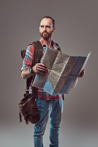 Free photo portrait of a handsome tattooed traveler in a flannel shirt with a backpack, holds a map, standing in a studio. isolated on a gray background.