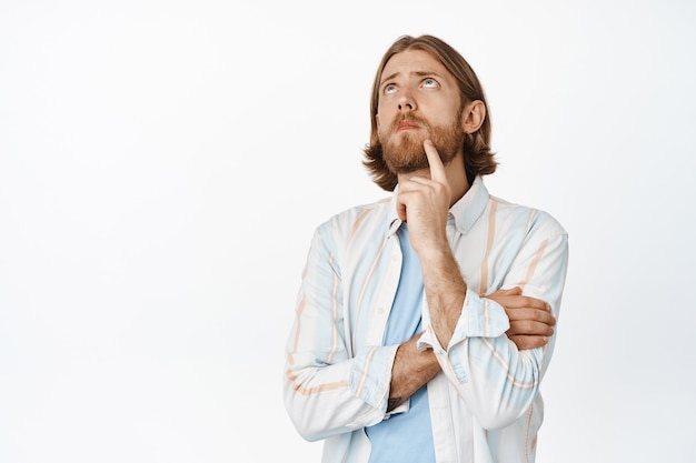 Portrait of handsome thoughtful blond man, touching his beard, frowning and looking up, thinking of something interesting, pondering, standing on white.