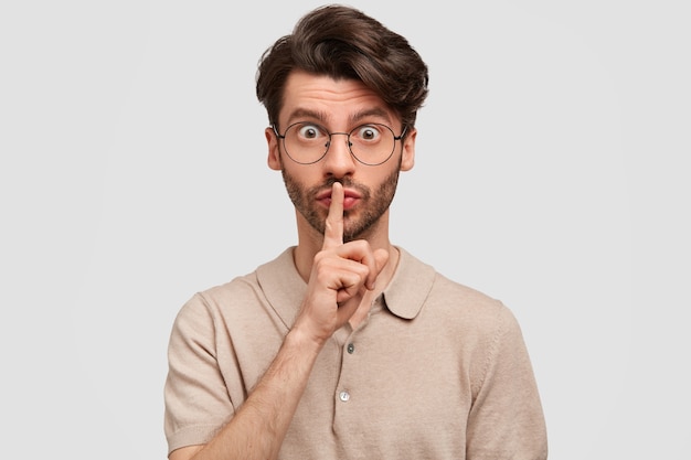 Portrait of handsome unshaven male keeps fore finger on lips