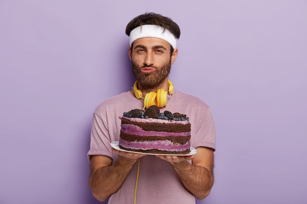 Free photo portrait of handsome unshaven man holds delicious cake on plate, dressed in casual wear, has good will not to eat dessert stands against purple wall, enjoys good taste. male with sweet dish