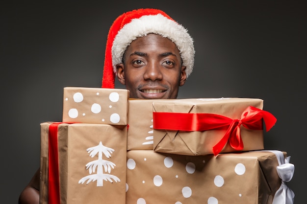 The portrait of handsome young black smiling man in Santa hat with gifts on dark.