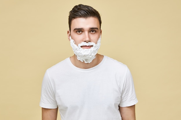 Portrait of handsome young Caucasian brunette man posing isolated with white shave foam applied on his face, preparing skin for shaving. Morning routine, masculinity, beauty and care concept