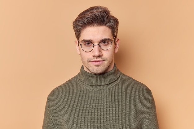 Free photo portrait of handsome young european man looks directly at camera with serious expression feels self confident wears round transparent glasses and casual sweater isolated over beige background.