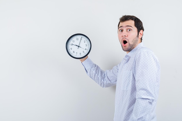 Portrait of handsome young man in white patterned shirt showing clock and worried about time isolated on white background