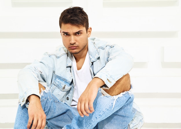 Free photo portrait of handsome young model man dressed in jeans clothes sitting near white textured wall