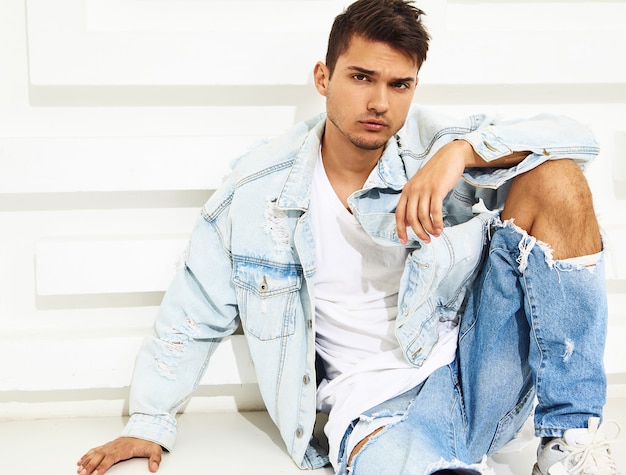 Portrait of handsome young model man dressed in jeans clothes sitting near white textured wall