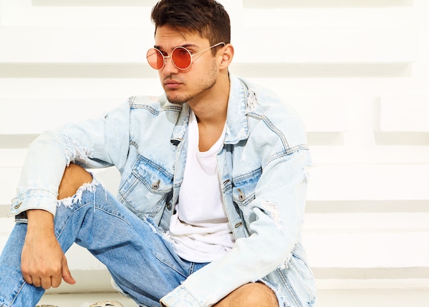 Free photo portrait of handsome young model man dressed in jeans clothes sitting near white textured wall