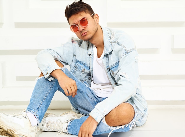 Portrait of handsome young model man dressed in jeans clothes sitting near white textured wall