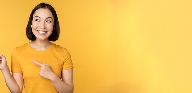 Portrait of happy asian girl pointing fingers and looking left smiling amazed checking out promo banner showing advertisement against yellow background