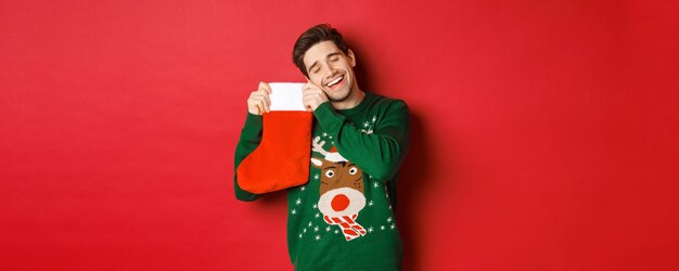 Portrait of happy attractive man in green sweater, looking delighted to receive gifts in christmas stocking, standing over red background.