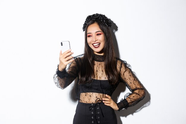 Portrait of happy beautiful asian woman in halloween costume smiling and looking at mobile phone screen, having video call, standing over white wall