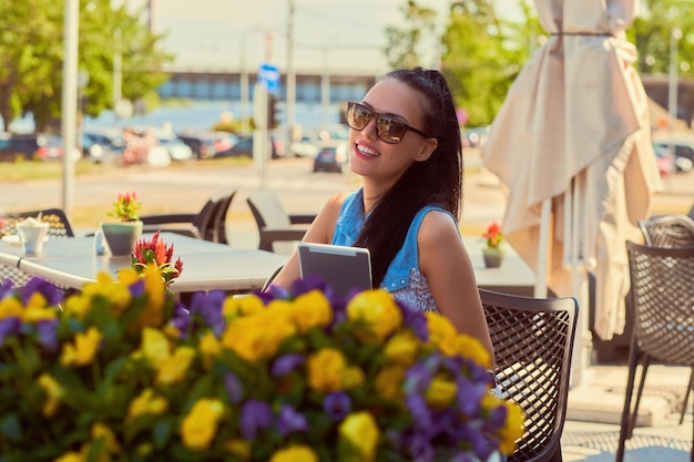 Portrait of a happy beautiful blogger girl wearing trendy clothes and sunglasses holds a tablet is enjoying summer day while sitting on a terrace in outdoors cafe, looking at the camera.