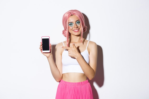 Free Photo portrait of happy beautiful female model in pink glamour wig and bright makeup, pointing finger at mobile phone screen, showing application or banner.