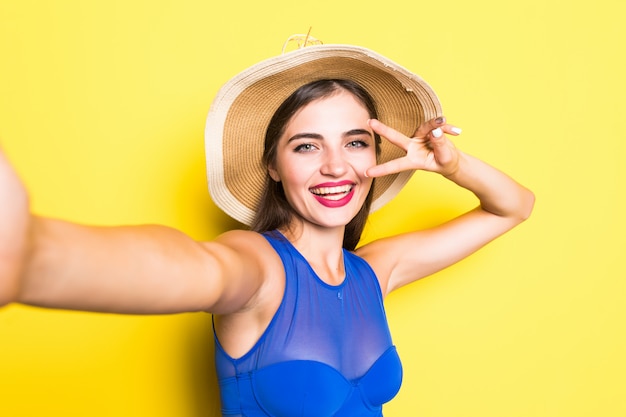 Portrait of happy beautiful young woman taking selfie on her phone and smiling, wearing swimsuit in straw hat on yellow wall