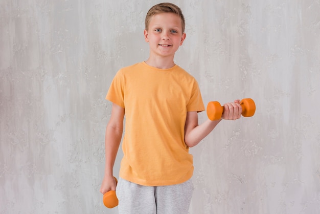 Portrait of a happy boy exercising with dumbbell standing in front of concrete wall