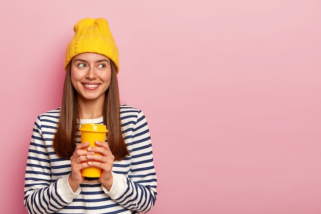 Portrait of happy brunette woman holds takeaway coffee, wears yellow hat and casual striped jumper, looks aside with smille, isolated over pink wall