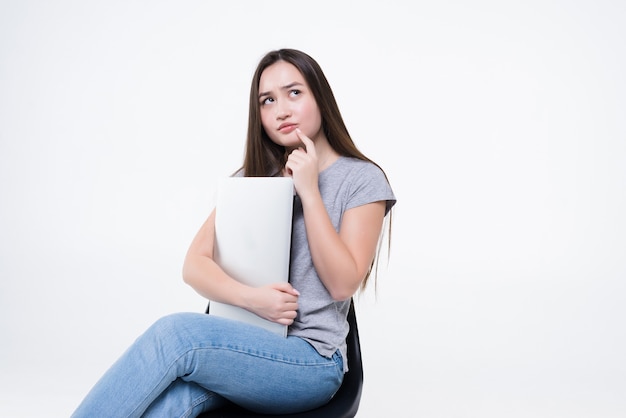 Portrait of a happy casual asian woman holding laptop computer while sitting on a chair over white wall