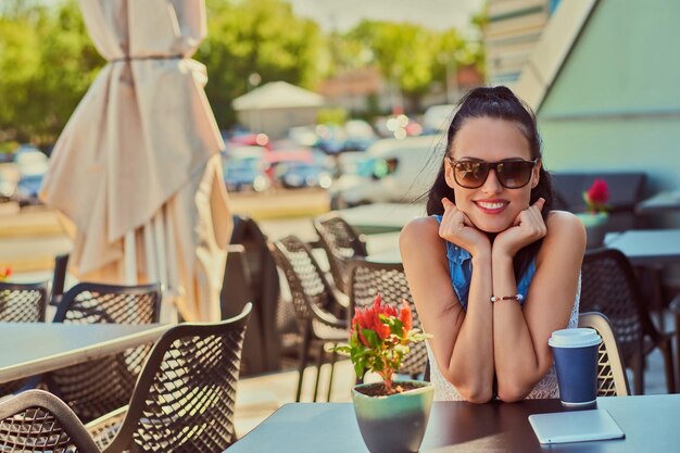 Portrait of a happy charming brunette girl wearing trendy clothes is enjoying summer day while sitting on a terrace in outdoors cafe, looking at the camera.