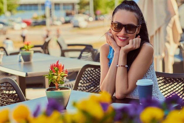 Portrait of a happy charming brunette girl wearing trendy clothes is enjoying summer day while sitting on a terrace in outdoors cafe, looking at the camera.