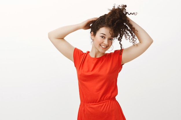 Portrait of happy charming caucasian woman in fashionable red dress, holding curly hair while combing it with both hands