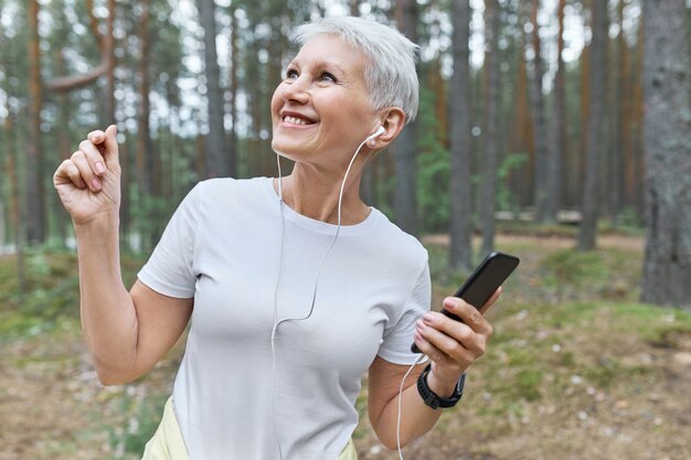 Portrait of happy cheerful mature woman in white t-shirt and earphones having fun outdoors