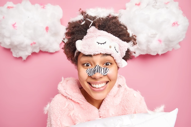 Portrait of happy curly haired young woman applies blackheads removal path on nose smiles broadly undergoes beauty skin care procedures dressed in pajama poses with pillow and flying feathers around