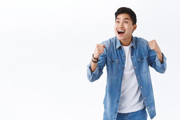 Portrait of happy energized young excited asian man watching sports game on tv in pub cheering for team scored goal smiling pleased clench fists in victory success gesture triumphing