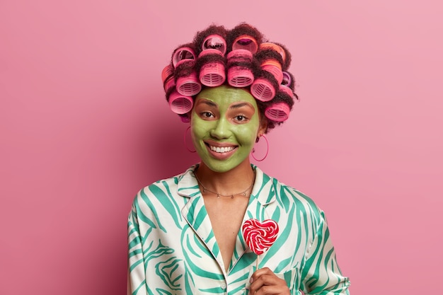 Portrait of happy ethnic woman wears green facial mask, smiles toothily, holds lollipop, makes curly hairstyle with rollers, wears casual domestic clothes, 