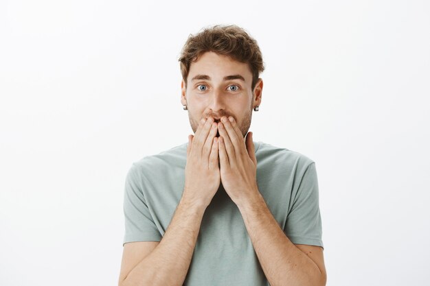 Portrait of happy european male model in earrings, smiling and covering mouth with palms, being amazed and pleased with great news