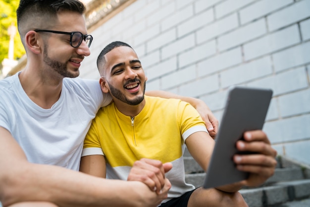 Portrait of happy gay couple spending time together and taking a selfie with digital tablet while sitting on stairs.