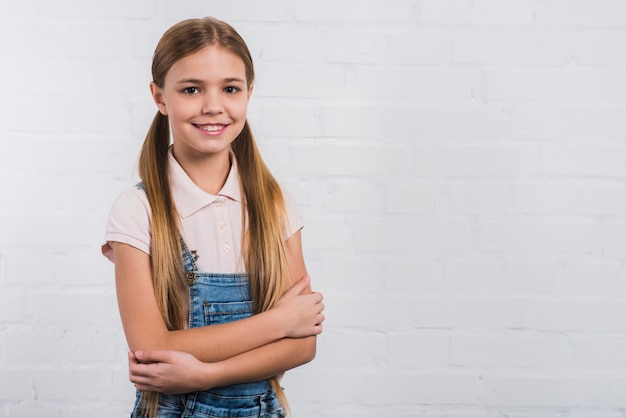 Free photo portrait of a happy girl with arm crossed looking to camera standing against white wall