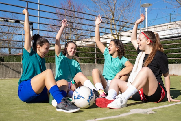 Free Photo portrait of happy girls talking on football field. four pretty caucasian girls in sportswear sitting on ground, raising hands to demonstrate belief in victory. healthy lifestyle and team sport concept