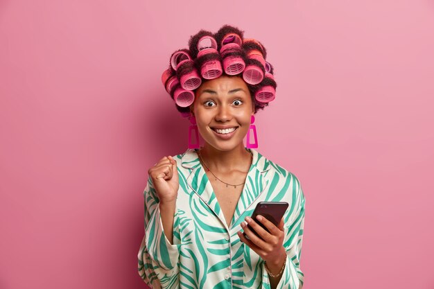 Portrait of happy housewife clenches fist and smiles broadly, dressed in casual domestic robe, makes hairstyle, applies hair rollers, waits for call, rejoices positive news isolated on pink wall