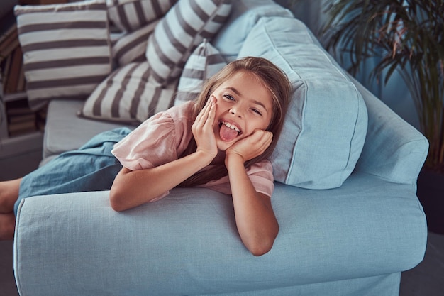 Free photo portrait of a happy little girl with long brown hair and piercing glance, shows tongue on the camera, lying on a sofa at home.