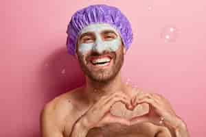 Free photo portrait of happy man with broad smile, enjoys washing and beauty treatments, has clay mask on face, stands topless