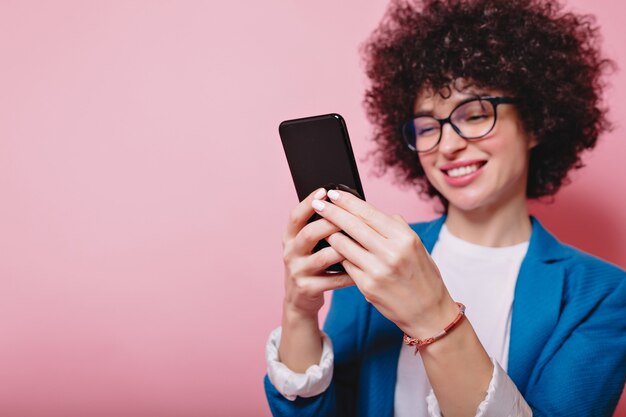 Portrait of happy modern woman with short hairstyle dressed blue jacket scrolling smartphone on pink with happy smile