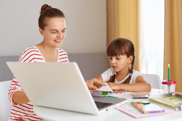 Portrait of happy positive female with daughter wearing casual attire, sitting at table against of window in living room, doing homework, mother helping child with online lesson.