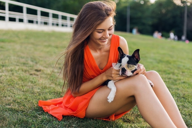 Portrait of happy pretty woman sitting on grass in summer park, holding boston terrier dog, smiling positive mood, wearing orange dress, trendy style, sunglasses, playing with pet