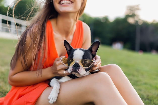 Portrait of happy pretty woman sitting on grass in summer park, holding boston terrier dog, smiling positive mood, wearing orange dress, trendy style, sunglasses, playing with pet