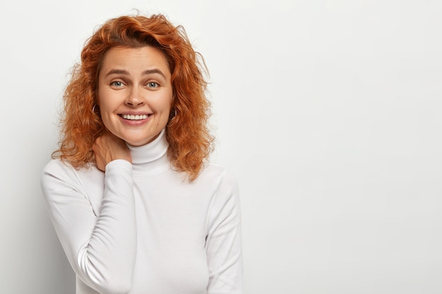 Free photo portrait of happy redhead curly woman feels enthusiastic and amused, touches neck gently, smiles gently , has nice friendly conversation, poses indoor against white wall