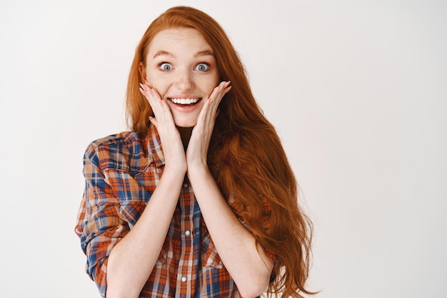 Free photo portrait of happy redhead girl hear amazing news looking surprised and happy touching cheeks and smiling at camera white background