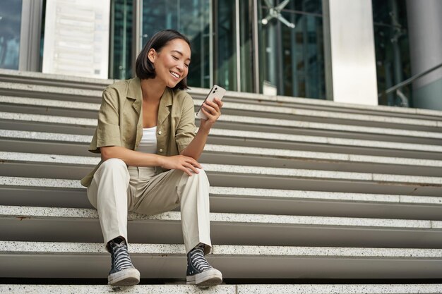 Portrait of happy smiling asian woman sitting outdoors near building using smartphone technology con